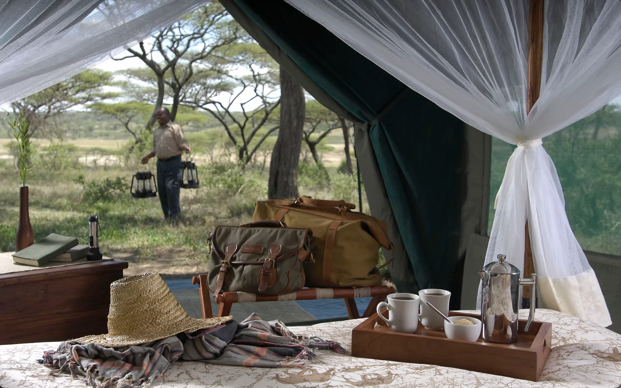 Morning coffee on the bed of a Serengeti camp tent, its mosquito net tied back, while a member of the camp team carries lanterns outside